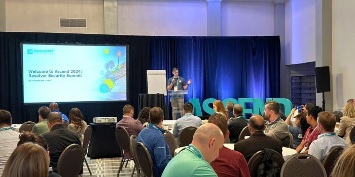 Reflecting on ascend 2024: resolver security summit 2 A speaker addressing the audience at ascend 2024, with a welcome slide on the screen and attendees seated in a conference room.