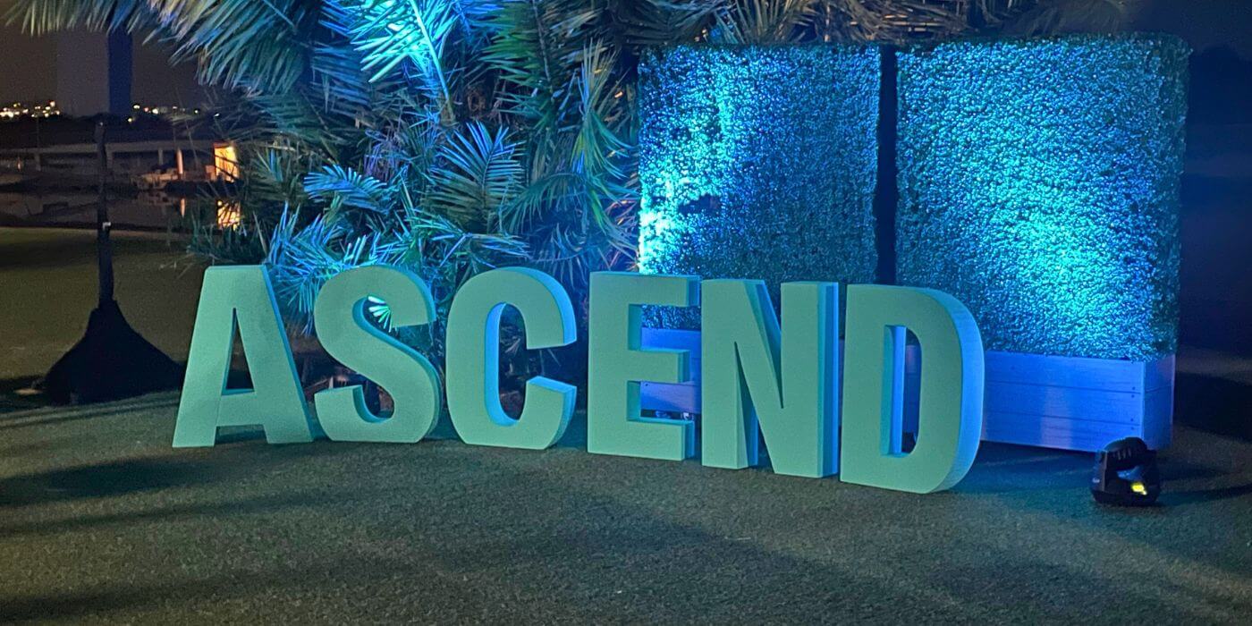 Large illuminated letters spelling 'ascend' at the ascend 2024 security summit, set against a backdrop of palm trees and a green hedge, with blue lighting creating a vibrant atmosphere.