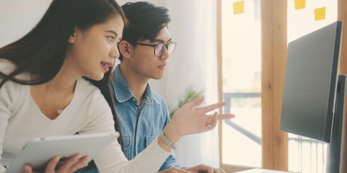 Woman holding a tablet leaning over a man sitting at a desk, and pointing at a computer monitor representing risk and compliance software