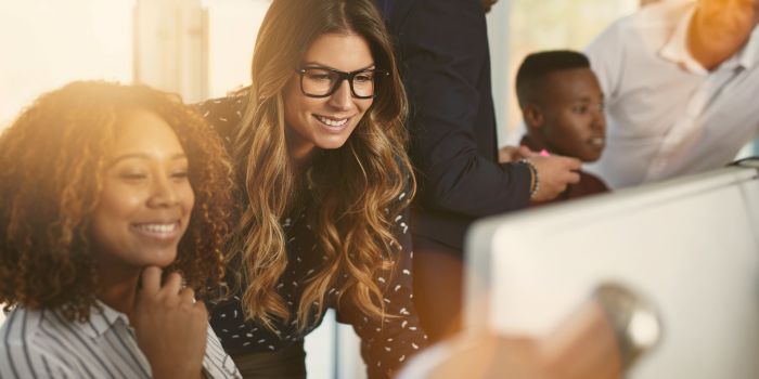 Two women, one standing the other sitting, in front a computer in the foreground, three men in the background, representing building a risk-aware culture