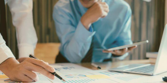 Woman holding pen on graphs in foreground, man watching with hand on his chin and open laptop in front of him representing risk mitigation strategies