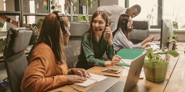 Two women wearing headsets with mics facing each other at a desk in the foreground; a man and woman at the end of the desk in background representing risks for operations teams
