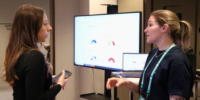 Two women in conversation at a tech demo during a conference, with data dashboards displayed on a monitor and laptop screen in the background.