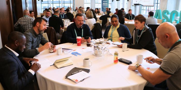 Attendees seated around a round table engaged in discussion during a conference session, with notebooks, drinks, and event lanyards visible; the large teal "ascend" letters can be seen in the background.