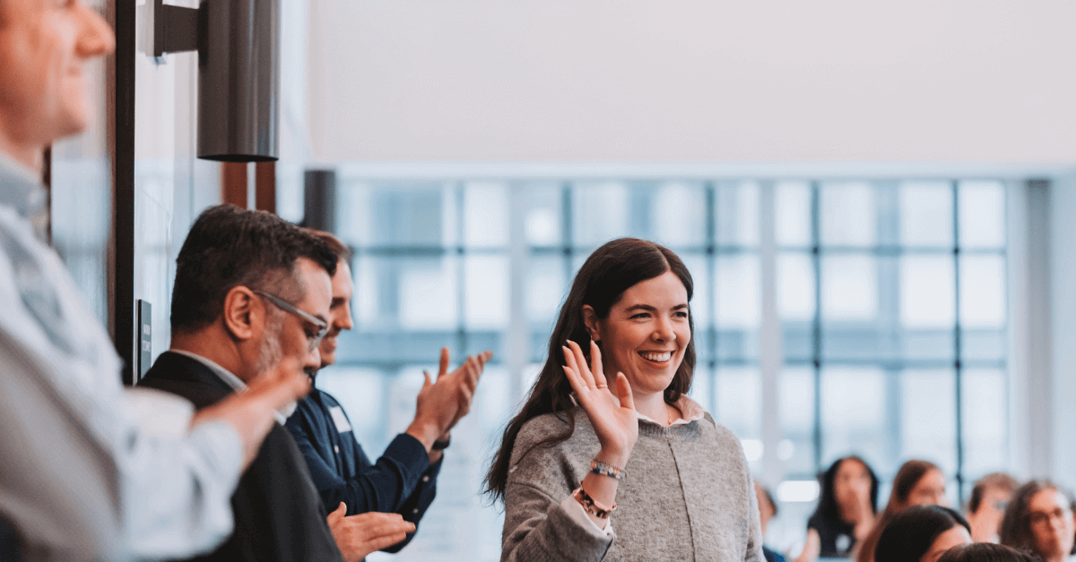 A smiling woman waving in greeting, surrounded by applauding colleagues at a networking event during grc day 2024