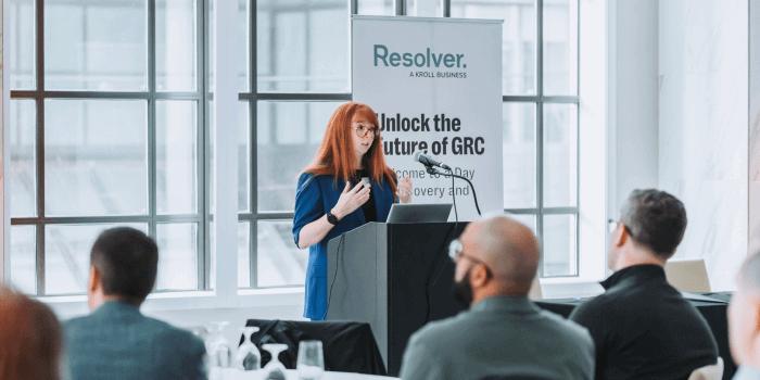 Ux research lead, sydney lamorea, at a podium for resolver's grc day, speaking to a crowd with a banner behind her reading "unlock the future of grc. "